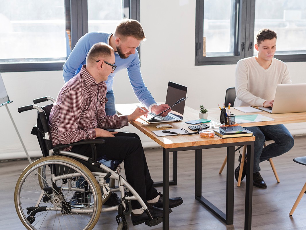 Two men working on a laptop together, one sitting in a wheelchair and the other standing beside him.